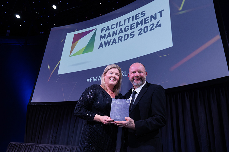 Two people dressed in black tie holding a square award. Behind them is a screen showing 'Facilities Management Awards 2024'